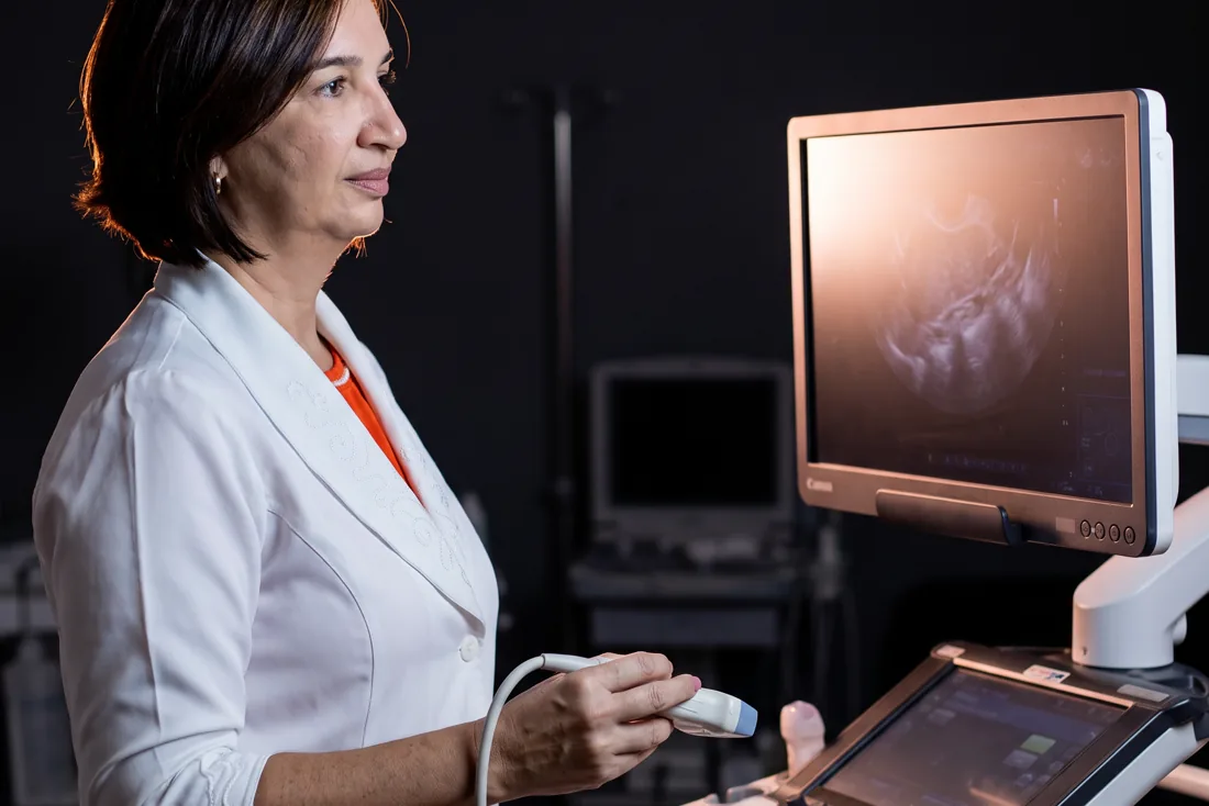 Dra. Daniella Prudente ao lado do aparelho de ultrassom, com o transdutor na mão, olhando para a imagem no monitor