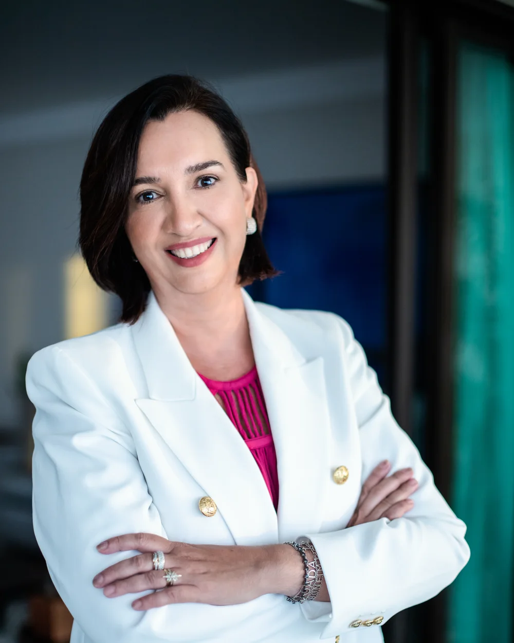 Dra. Daniella Prudente de blazer branco, braços cruzados, sorrindo, em ambiente escuro